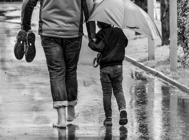 Black and white photo, dad and son are walking through the park in the rain, the dad with bare feet