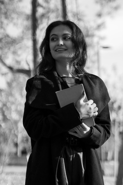 Black and white photo of a girl in a black coat walking with a book in her hands
