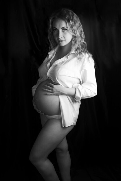 Black and white photo of a pregnant woman in the studio on a black background, standing half-turn knee-deep. In a white shirt and underwear, hands on a bare belly.One knee bent. Looking at the camera