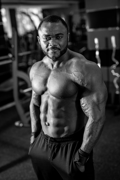 Foto in bianco e nero dell'uomo forte con un tipo di corpo muscoloso in posa nel moderno palazzetto dello sport uomo multirazziale che guarda la telecamera con un sorriso leggero