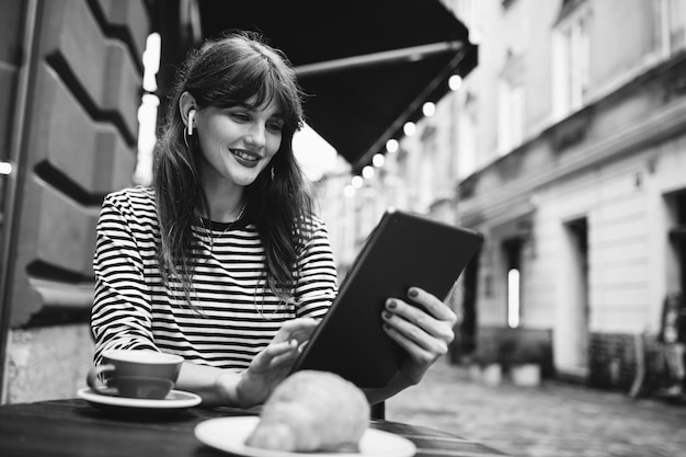 Black and white photo of  woman drinking a coffee with digital tablet and earphones on cafe terrace