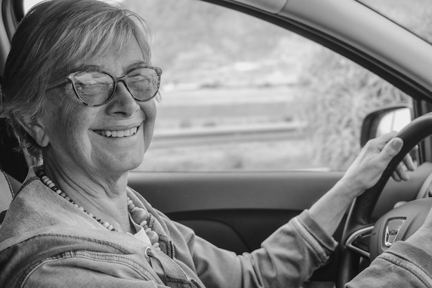 Foto ritratto in bianco e nero di donna anziana felice attraente con gli occhiali che guida l'auto con le mani sul volante