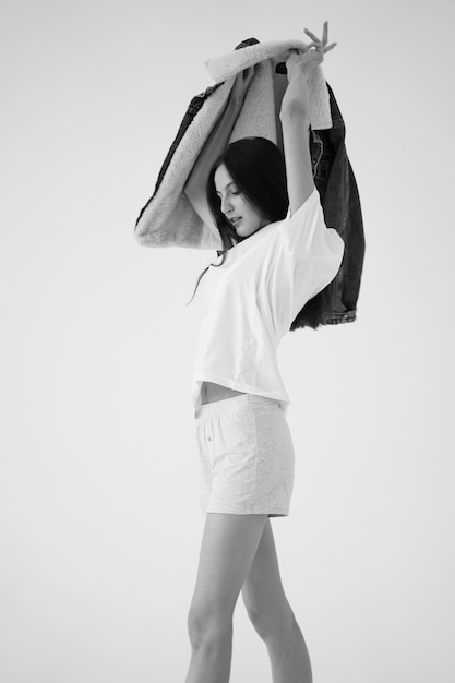 Black and white portrait A beautiful young girl in a white Tshirt and short white shorts puts on a denim jacket on a white background