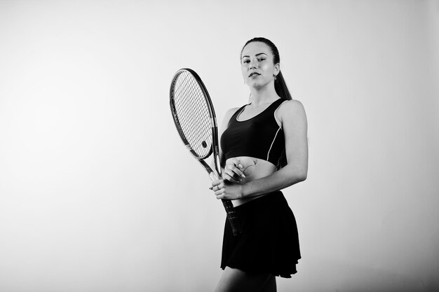 Black and white portrait of beautiful young woman player in sports clothes holding tennis racket while standing against white background