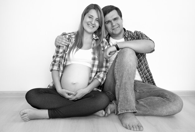 Черно-белый портрет счастливой беременной пары, сидящей на полу и смотрящей друг на друга