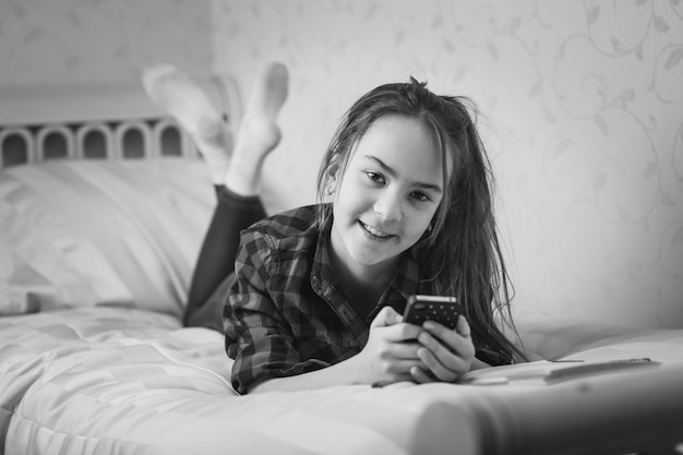 Black and white portrait of happy teenage girl lying on bed and typing message