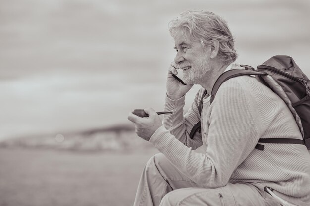 Black and white portrait of senior man sitting on the cliff in sea excursion smoking pipe looking at the horizon while talking on mobile phone