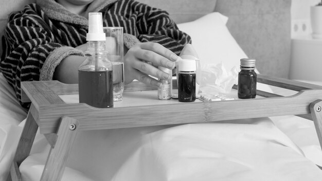 Black and white portrait of sick woman taking medicines fom wooden tray in bed.