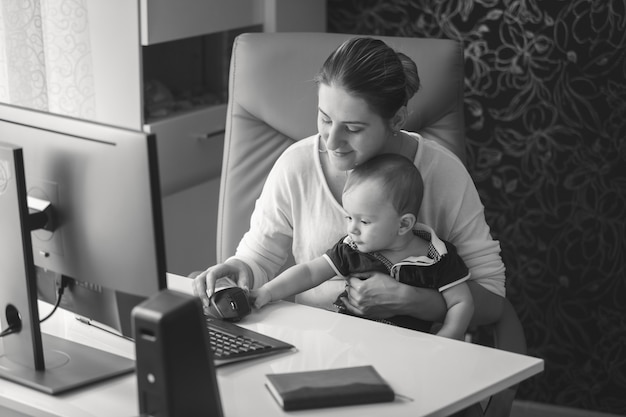 Черно-белый портрет улыбающейся молодой матери, сидящей в офисе с ребенком