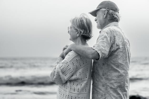 Черно-белый портрет двух красивых и счастливых пожилых людей или пенсионеров, обнимающихся на пляже в свете заката, пожилая улыбающаяся пожилая пара на открытом воздухе, наслаждающаяся отдыхом вместе