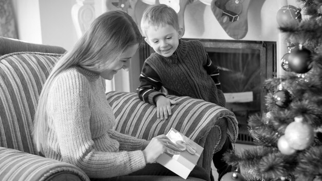 Ritratto in bianco e nero di una giovane madre con il figlioletto che apre una scatola con regali e regali di natale