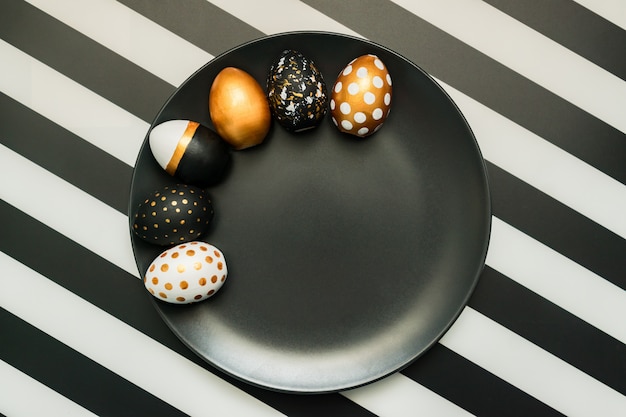 Photo black and white striped background with white, black and golden decorated eggs on black plate. trendy easter flat lay.