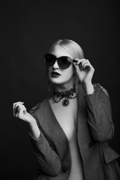 Black and white studio portrait of a stylish blond woman wears sunglasses and jacket on a naked body