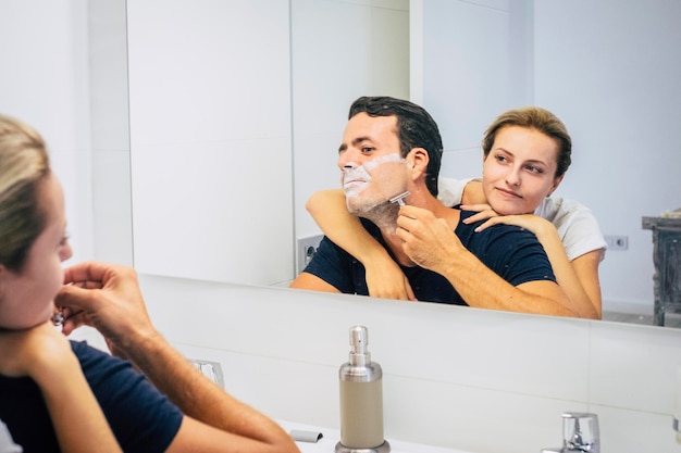 Black and white young couple having fun in the bathroom at home\
man shaving beard in morning routine and girl smiling concept of\
living together in relationship happy people indoor leisure