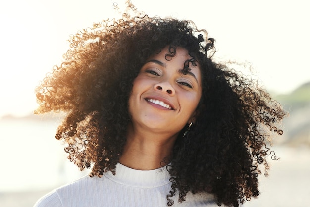 Black woman afro and wind in hair outdoor with a smile and portrait at beach for vacation or freedom Face of happy young model person in nature for peace travel and time to relax on sunset holiday