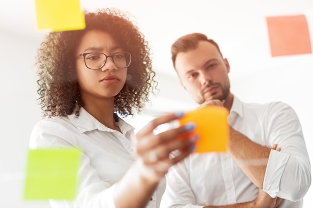 Foto donna di colore allegando una nota adesiva sul vetro mentre si riflette sulla strategia creativa durante la riunione in ufficio