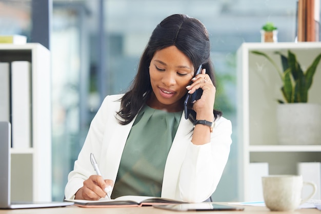 Black woman business and phone call while writing in notebook journal and planner at office desk Female entrepreneur talking on smartphone while planning notes for agenda schedule and information