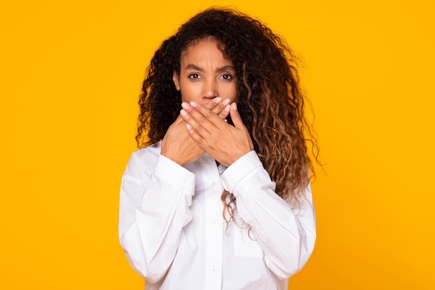 Black woman covering mouth with hands isolated on yellow background