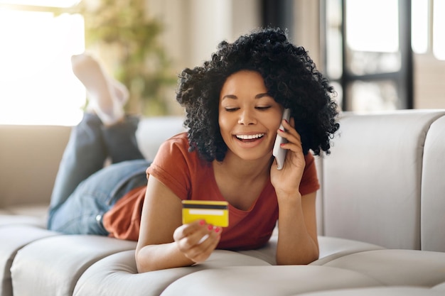 Black woman holding credit card and talking on mobile phone at home