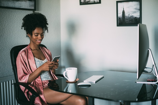 Donna di colore a casa che lavora con il computer, lo smartphone e il caffè di mattina