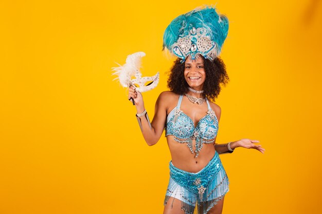 Black woman queen of brazilian samba school with blue carnival clothes and crown of feathers holding mask welcome