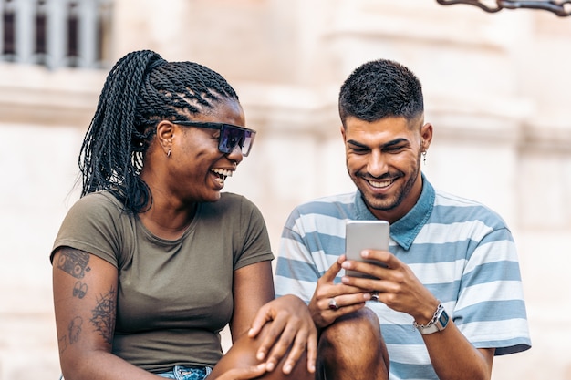 Black woman sitting next to a caucasian man laughing while looking at the mobile sitting outdoors