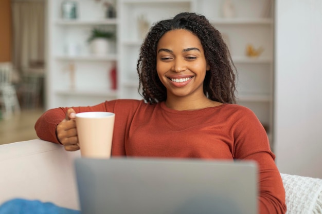 Black woman sitting on couch with coffee cup looking at laptop