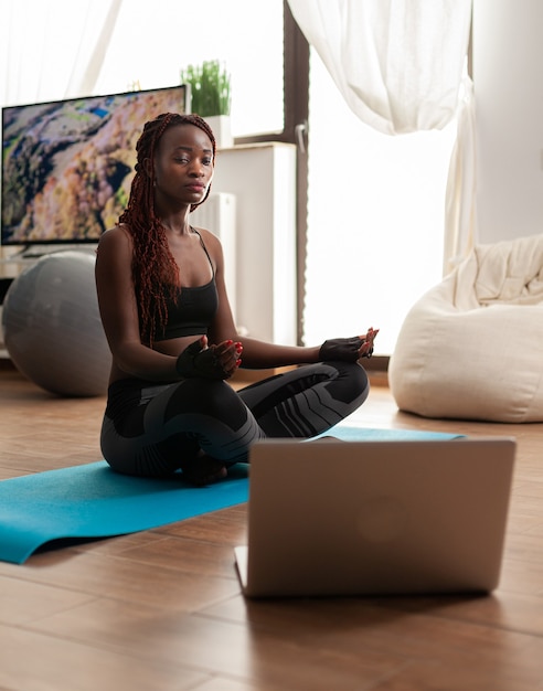 健康的なライフスタイルのために禅を瞑想する穏やかな調和を練習し、蓮華座でリラックスして、ヨガマットに座っている黒人女性。オンライントレーニング中にインストラクターの指示を聞く。