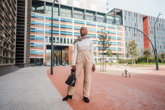 Black woman in smart casual wear and hijab standing near skyscraper