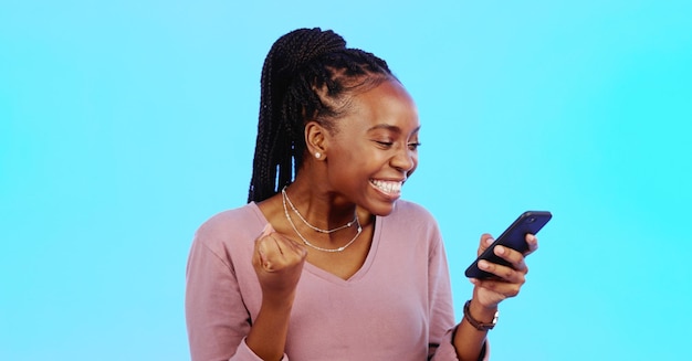 Photo black woman smartphone and fist for celebration lucky winner and achievement against a blue studio background african american female happy lady and cellphone with email social media and victory