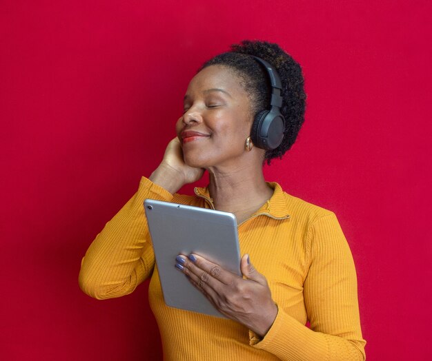 Black woman smiling and holding on a tablet, doing a gesture and listening song with  a yellow shirt in a red background