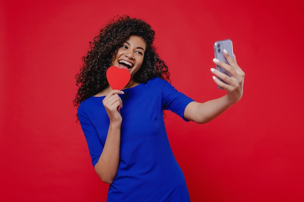 Black woman smiling and making selfie with heart shaped valentine card on red wall