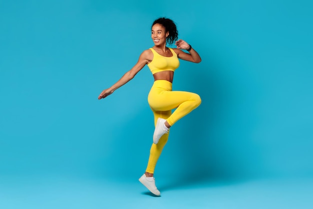 Black woman in sportswear jumping doing exercise on blue backdrop