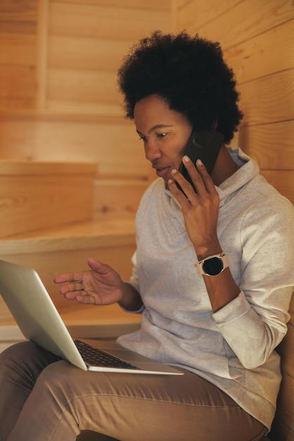 Black woman talking on a smartphone while using a laptop during holiday in a cozy wooden house in a forest