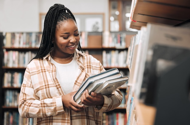 Black woman university student and library with books for education learning and knowledge at campus Happy smile and gen z college student at bookshelf for studying academic research and reading