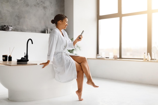 Photo black woman in white bathrobe laughing while texting on her smartphone