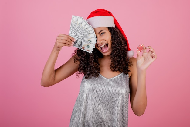 Black woman with casino poker chip and cash money wearing holiday santa hat over pink