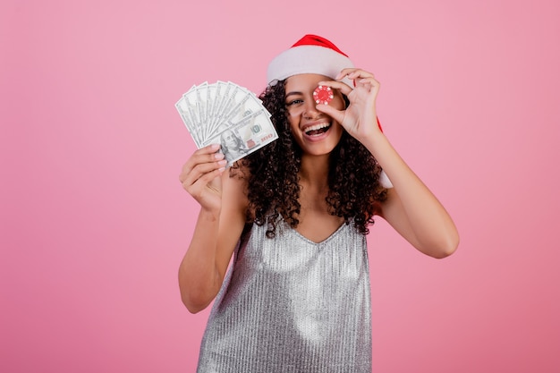 Black woman with casino poker chip and cash money wearing holiday santa hat over pink