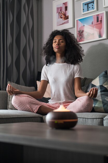 Black woman with curly hair sitting on sofa in Lotus pose and meditating