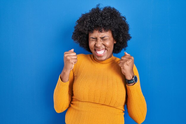 Black woman with curly hair standing over blue background excited for success with arms raised and eyes closed celebrating victory smiling winner concept