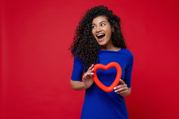 Black woman with heart shape smiling and wearing blue dress on red wall