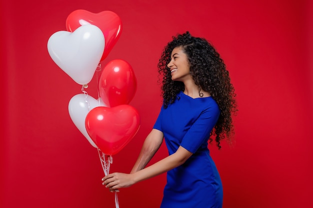 Black woman with heart shaped colorful balloons wearing blue dress on red wall