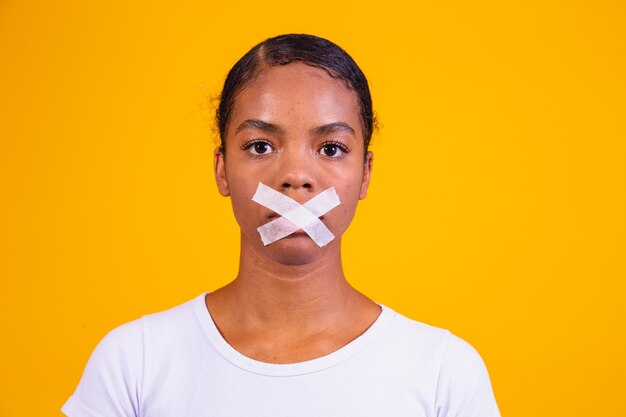 Black woman on yellow background with her mouth closed in silence. Concept of prejudice, abuse and racism