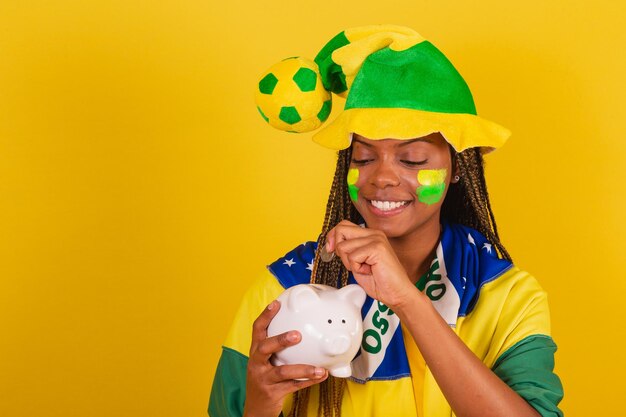 Black woman young brazilian soccer fan holding piggy bank and\
coin concept of finance economy