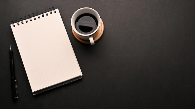 Black workspace table with a cup of coffee an empty spiral notepad and copy space