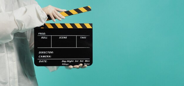 검은색과 노란색 클래퍼보드 또는 영화 슬레이트우먼은 민트 녹색 배경에 PPE 정장을 입습니다.
