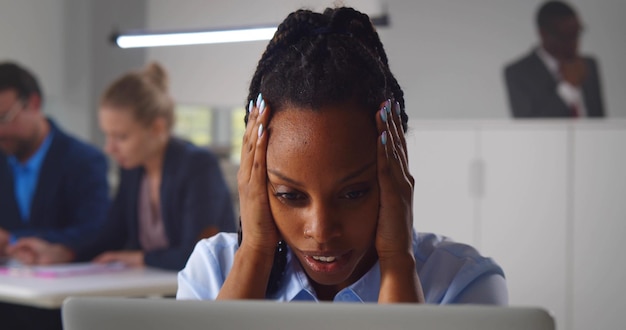 Black young businesswoman sitting at desk with laptop exhausted from online work in office