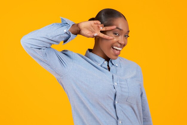 Black young woman gesturing victory sign near face yellow backdrop