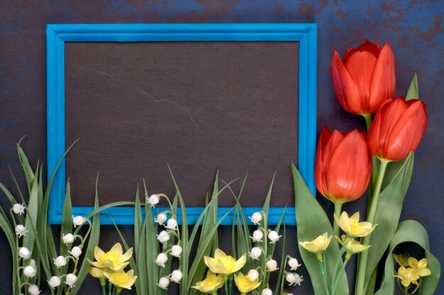 Blackboard in blue frame with red tulips and lily of the valley flowers on dark 