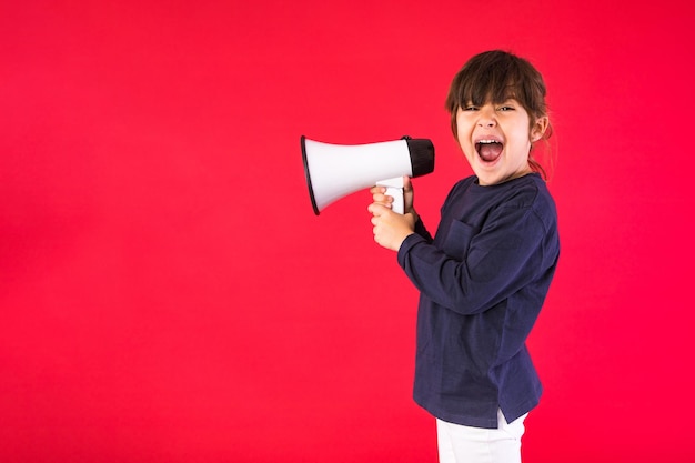 Blackhaired girl in a blue sweater and white pants shouting through a white megaphone on a red background